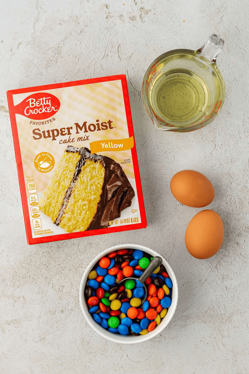 A box of Betty Crocker Super Moist yellow cake mix, two eggs, a glass measuring cup of oil, and a bowl of colorful M&M candy ready to make delicious cake mix cookies on a light-colored surface.