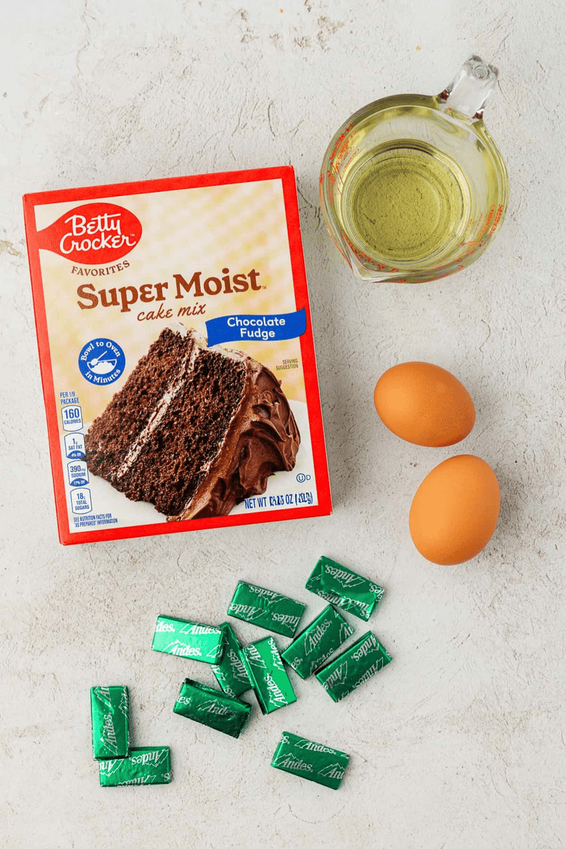 A box of Betty Crocker Super Moist chocolate fudge cake mix, a glass measuring cup with oil, two brown eggs, and several unwrapped Andes mint chocolates—perfect for making delicious cake mix cookies—are arranged on a light surface.