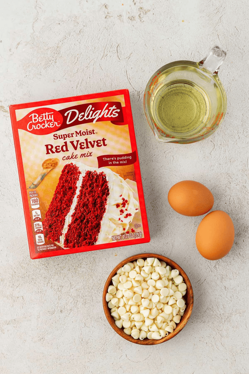 A box of Betty Crocker red velvet cake mix, a glass measuring cup with oil, two brown eggs, and a wooden bowl filled with white chocolate chips—everything you need to bake irresistible red velvet cake mix cookies on a light countertop.