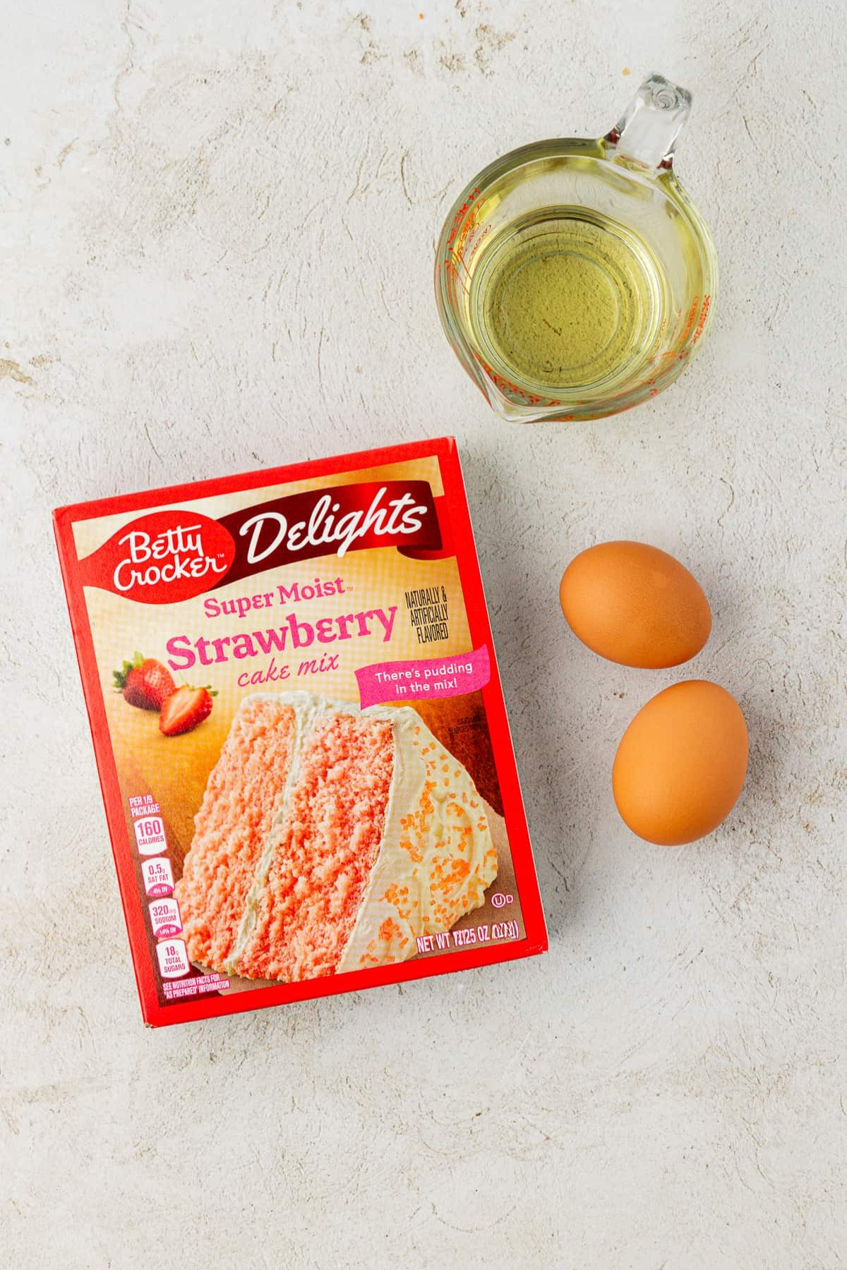 A box of Betty Crocker Super Moist Strawberry cake mix, two brown eggs, and a glass measuring cup with oil are arranged on a light-colored surface—perfect for making easy strawberry cake mix cookies.