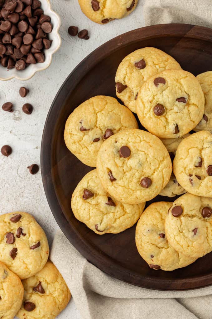 chocolate chip cake mix cookies on a brown plate on top of a tan linen towel, surrounded by sprinkled chocolate chips, more cookies and a small white bowl of chocolate chips