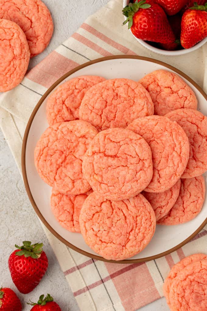 Strawberry cake mix cookies piled on top of a white plate with a brown rim, sitting on top of a tan and pink kitchen towel and surrounded by more cookies, strawberries and a small white bowl of strawberries