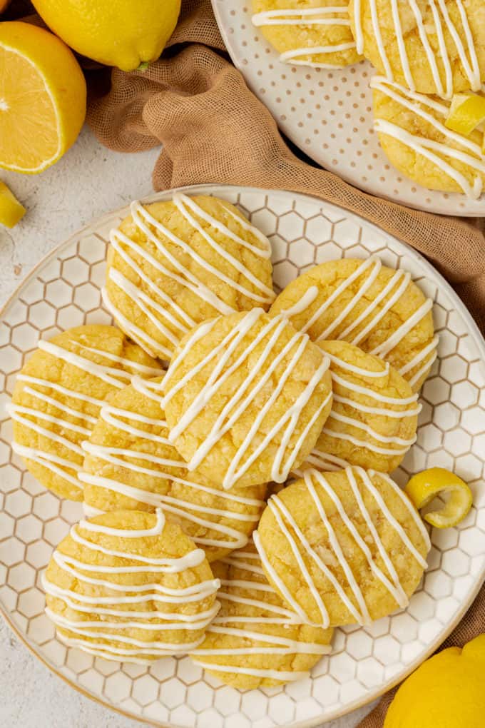 lemon cake mix cookies on a honeycomb plate surrounded by tan cloth, lemons and another plate of cookies in the background