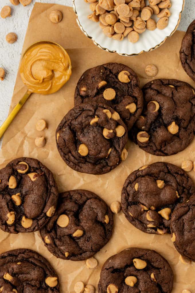 chocolate peanut butter cake mix cookies on brown paper with a spoon of peanut butter, a white bowl of peanut butter chips and more peanut butter chips sprinkled around