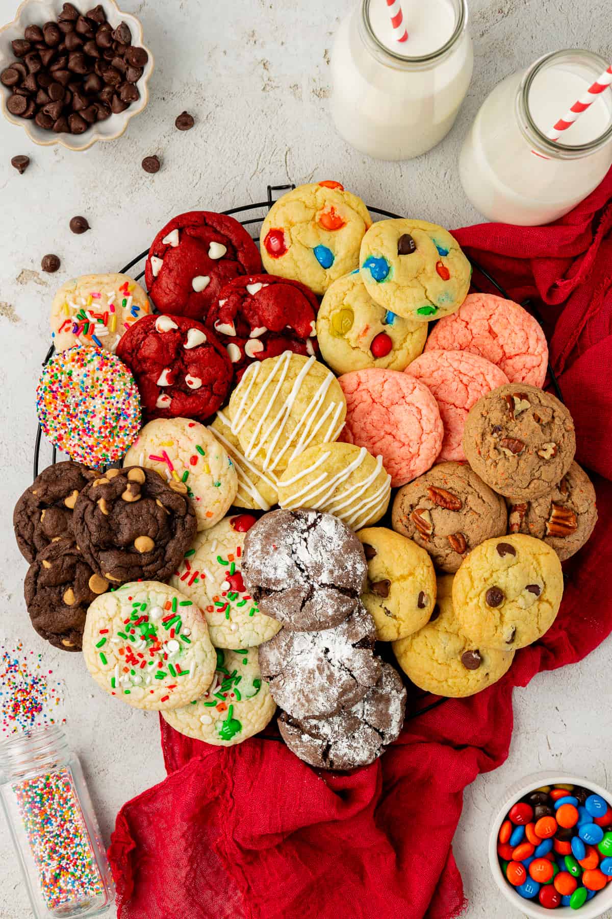 an assortment of cake mix cookies in different flavors in a cirlcle on a wire rack on top of a red towel with sprinkles, M&ms, chocolate chips and two glasses of milk with red and white straws all around