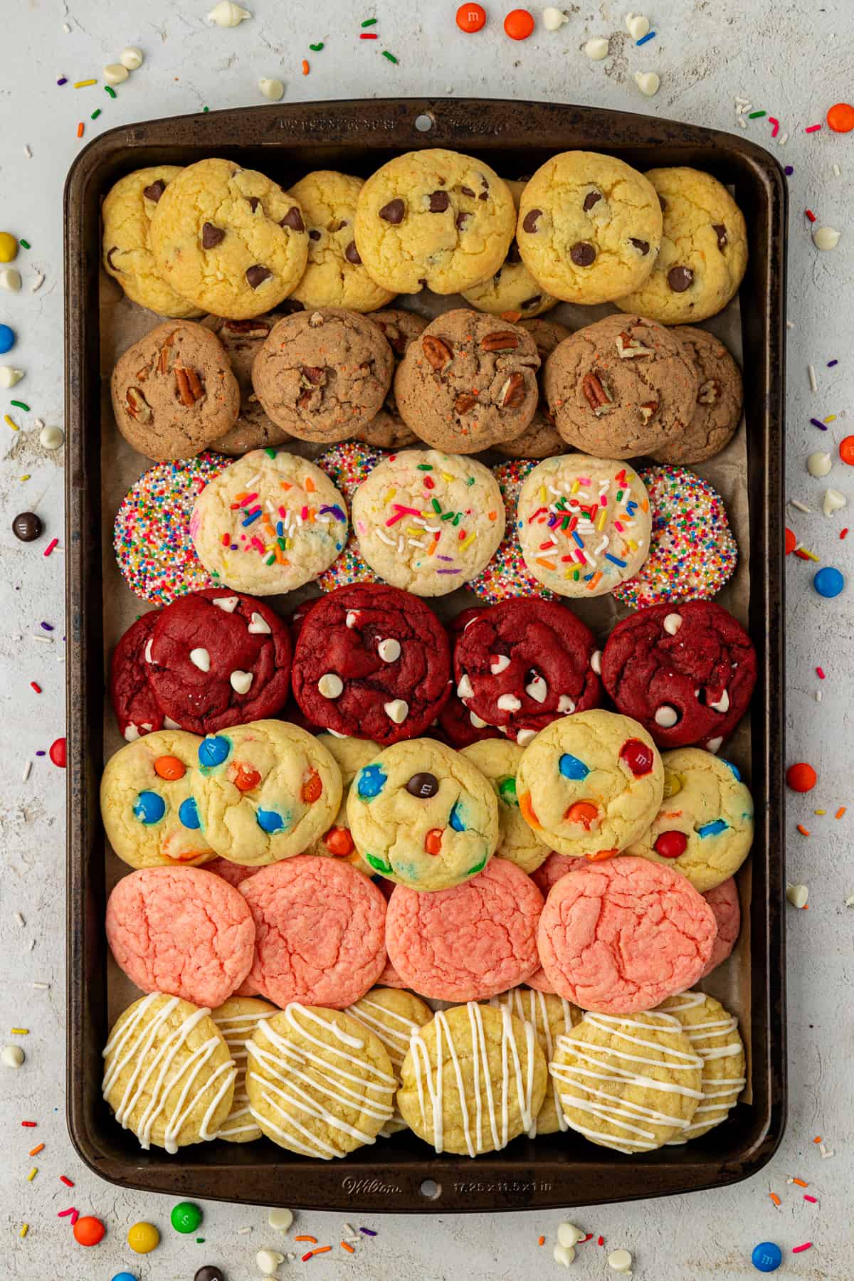 rows of assorted cake mix cookies stacked in a couple of layers on top of a baking sheet surrounded by m&ms, reese pieces and sprinkles