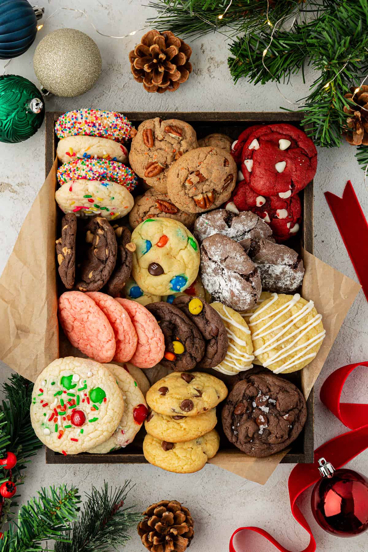 a box full of assorted cake mix cookies flavors with brown paper surrounded by christmas ornaments, red ribbon, pine cones and christmas tree pieces