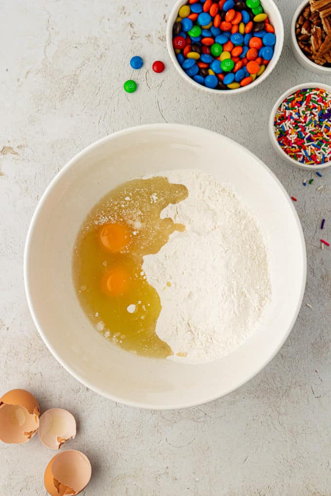 eggs and dry ingredients for cake mix cookies in a white bowl surrounded by egg shells, a bowl of sprinkles, a bowl of m&ms and a bowl of nuts
