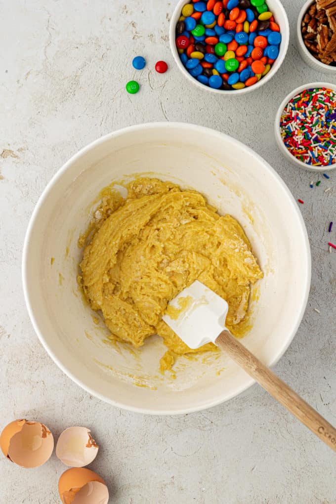 cake mix cookie batter in a white bowl with a white and wooden spatula surrounded by egg shells, a bowl of sprinkles, a bowl of m&ms and a bowl of nuts