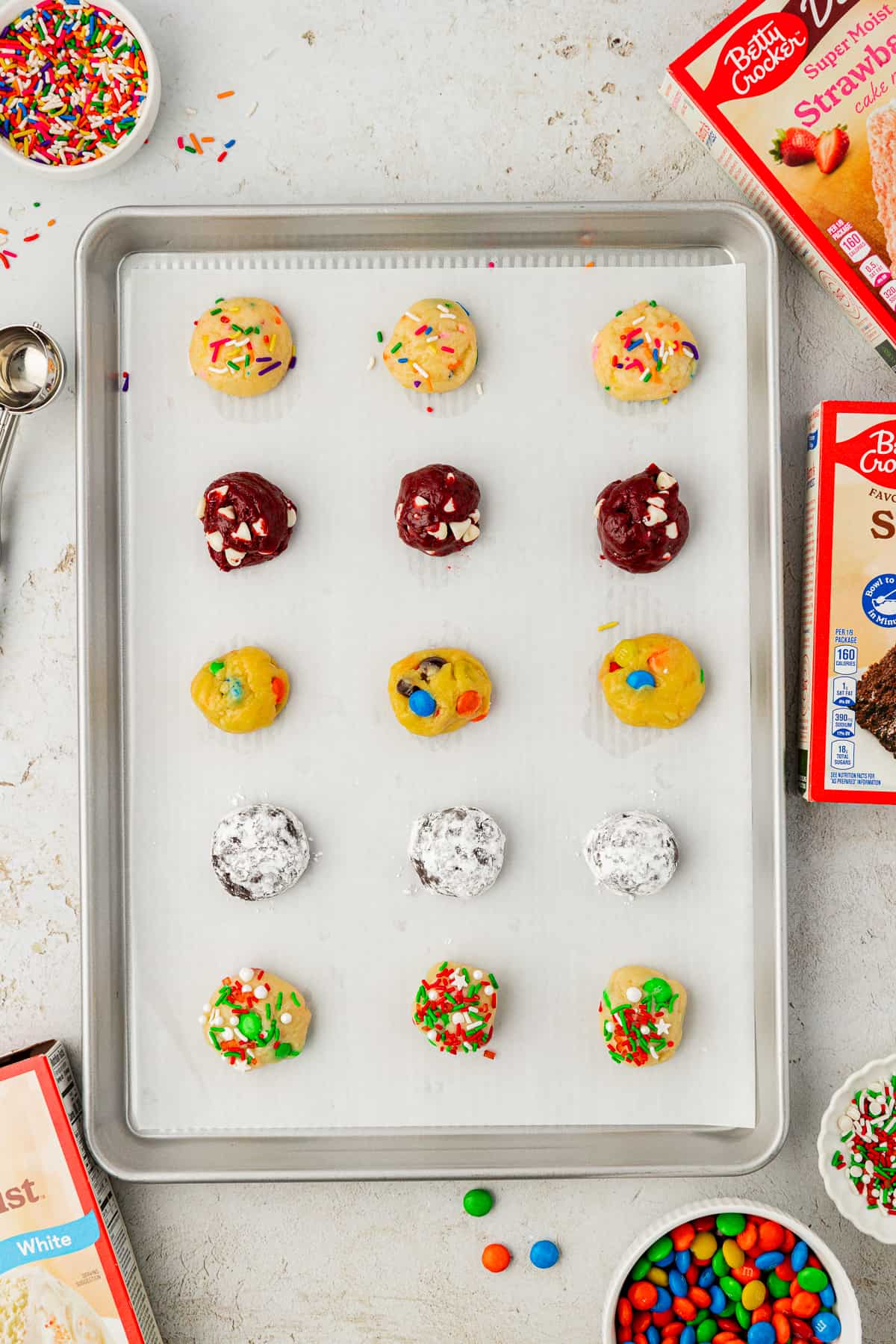 cookie dough balls of assorted flavors of cake mix cookies in rows on a baking sheet lined with parchment paper with boxed cake mix, M&Ms and sprinkles around it