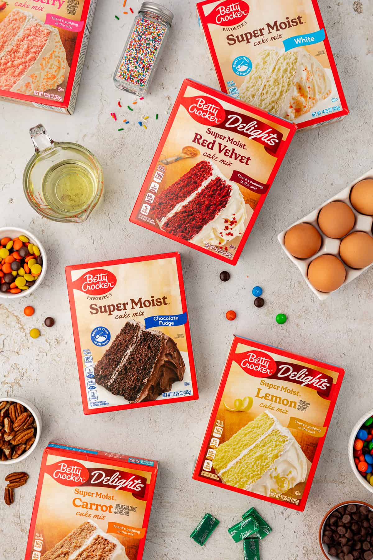 ingredients for cake mix cookies including vegetable oil, strawberry cake mix, white cake mix, red velvet cake mix, chocolate cake mix, lemon cake mix, eggs, m&ms, nuts, reese's pieces, sprinkles and andes mints