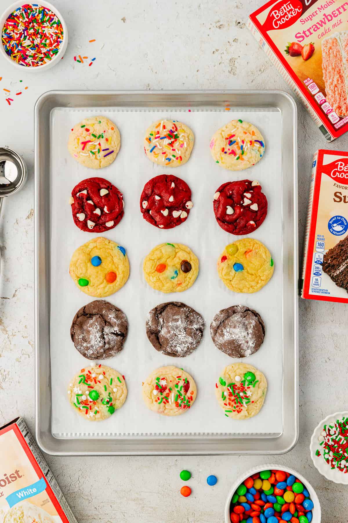 assorted flavors of cake mix cookies in rows on a baking sheet lined with parchment paper surrounded by boxes of cake mix, sprinkles, M&Ms and a cookie scoop
