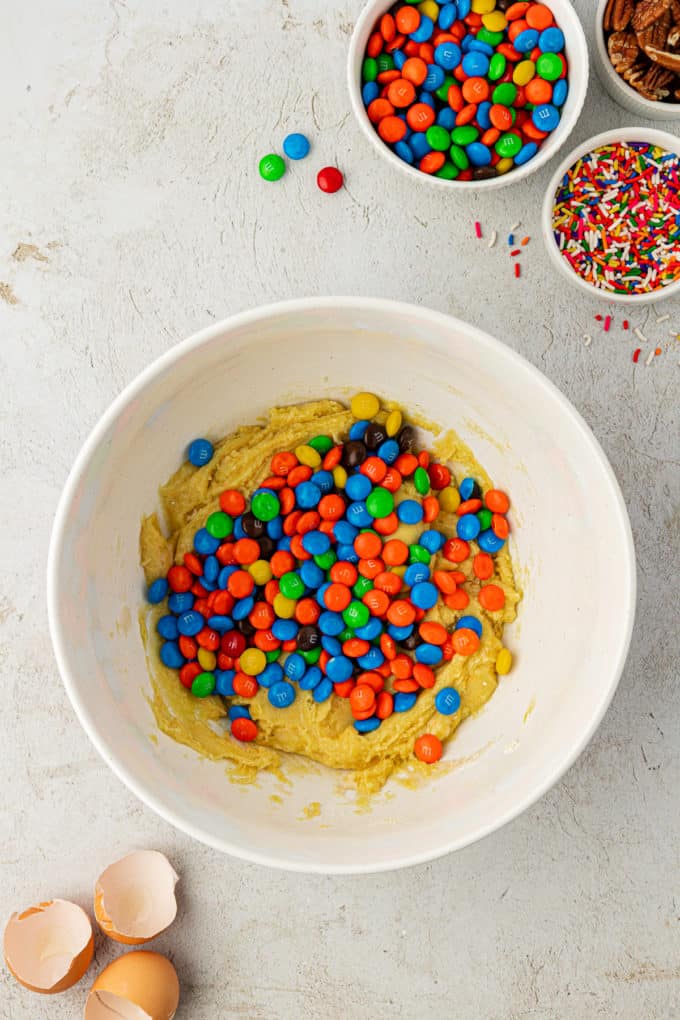 cake mix cookie batter in a white bowl with a pile of M&Ms on top, surrounded by egg shells, a bowl of sprinkles, a bowl of m&ms and a bowl of nuts
