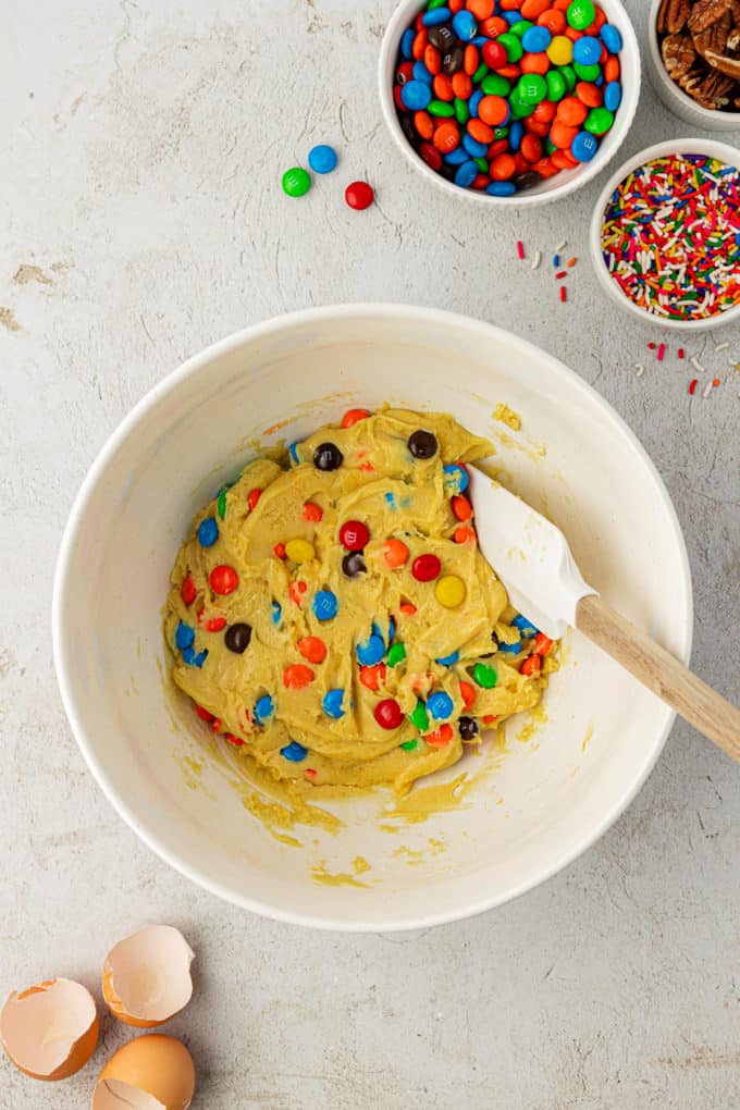 cake mix cookie batter in a white bowl with with M&Ms mixed in with a white and wooden spatula surrounded by a bowl of sprinkles, a bowl of m&ms and a bowl of nuts