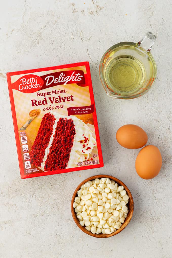 ingredients for red velvet cake mix cookies including a box of red velvet cake mix, two eggs, a wooden bowl of white chocolate chips and vegetable oil