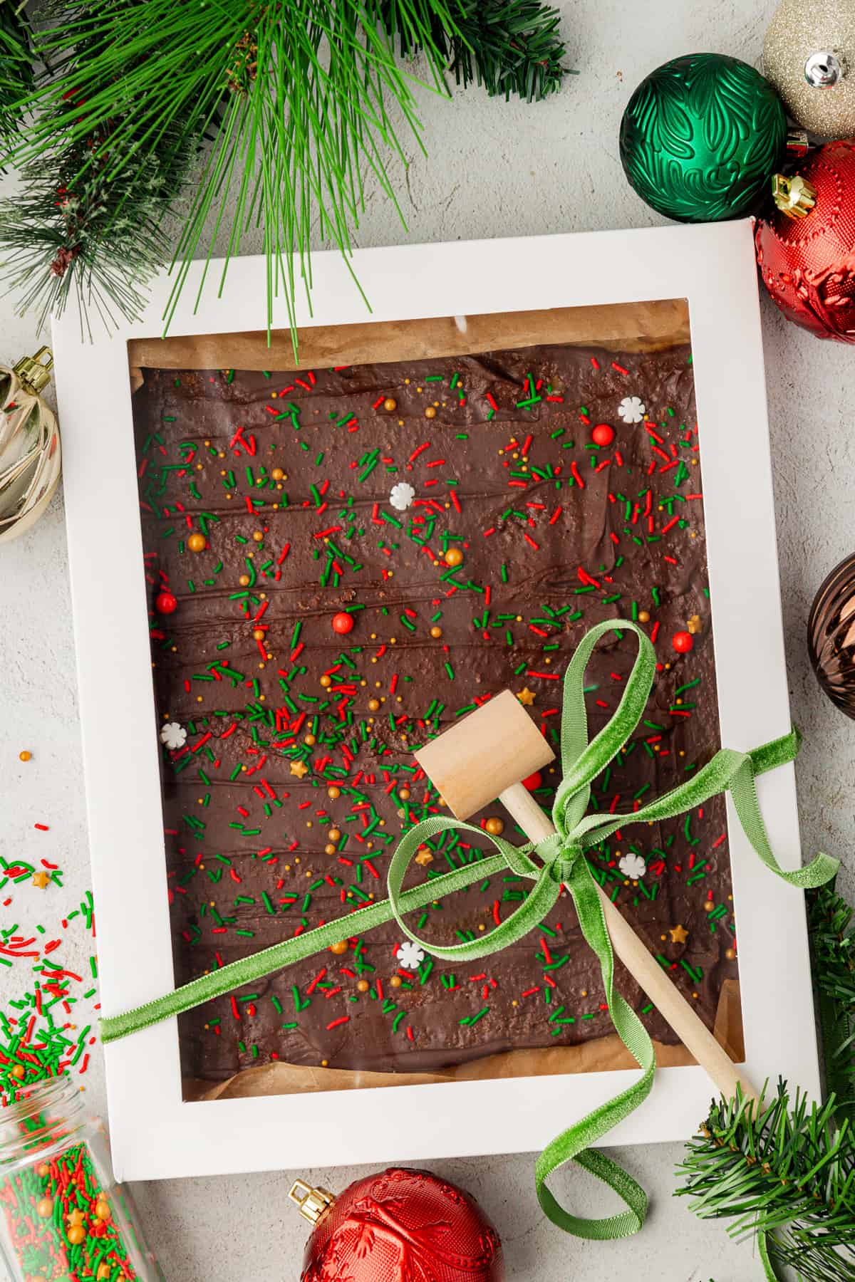 a large white box with a see-through top full of christmas crack candy with a green ribbon tied around it and a wooden mallet tied into the ribbon, with sprinkles, christmas ornaments and pine stems around