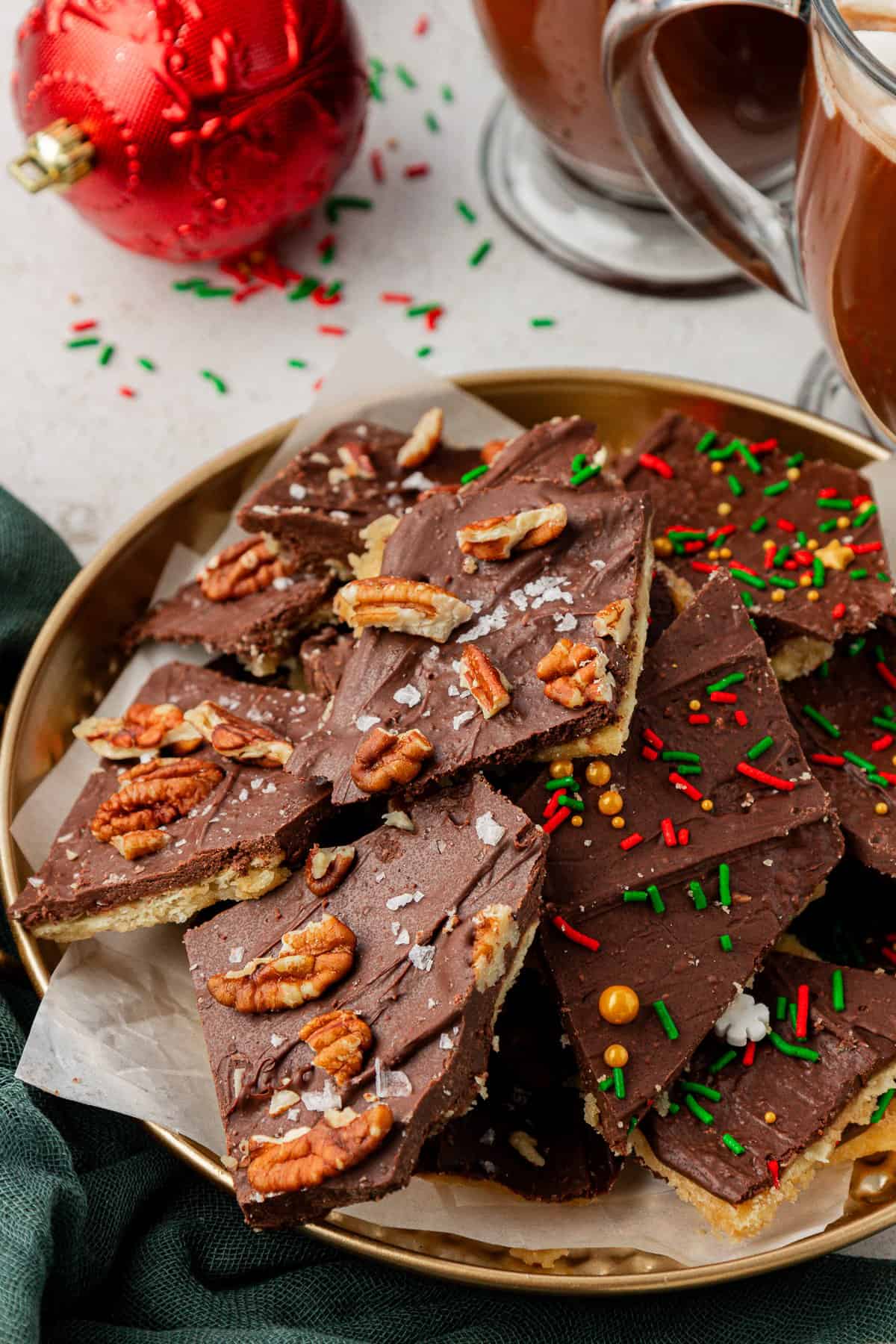 a gold plate with christmas crack on it, half with pecans and half with sprinkles, on a white surface with a green towel, a red christmas ornament, green and red sprinkles around and two glass mugs
