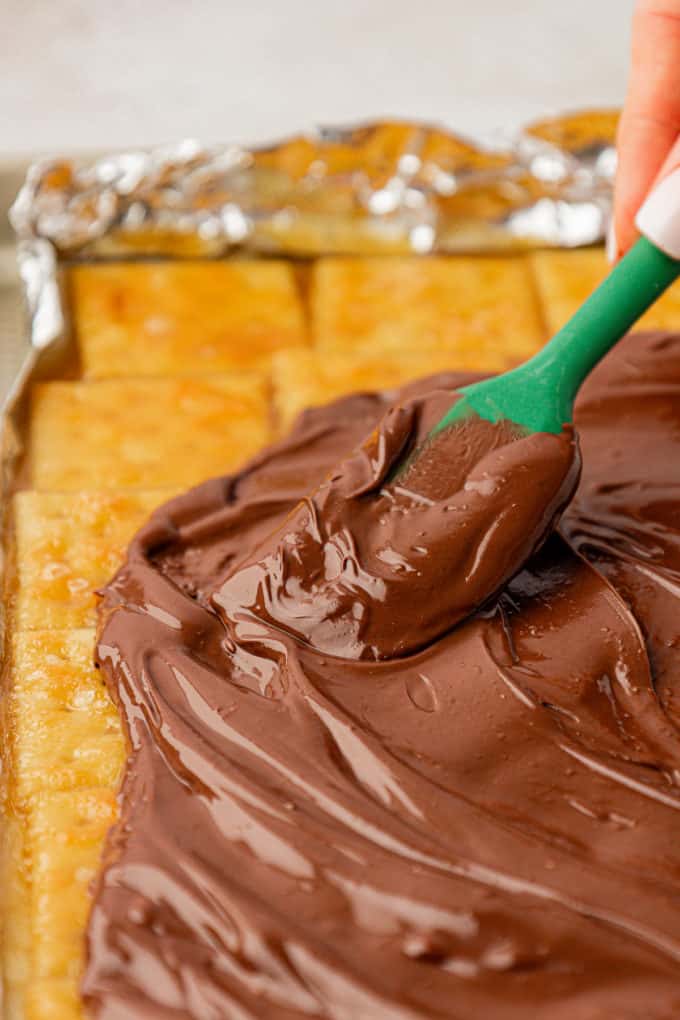 Melted chocolate being spread with a green spatula over a single layer of saltine crackers that have been cokked with a brown sugar butter mixture of top of them