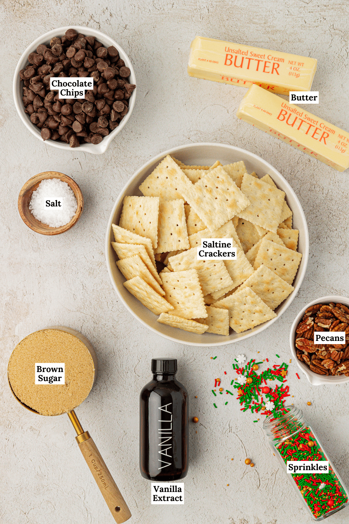 overhead view of ingredients for christmas crack including a bowl of saltine crackers, a measuring cup of brown sugar, a bowl of semi-sweet chocolate chips, a small bowl of sea salt, a bottle of sprinkles laying on its side with some spilled out, two sticks of butter, and a small bowl of pecans