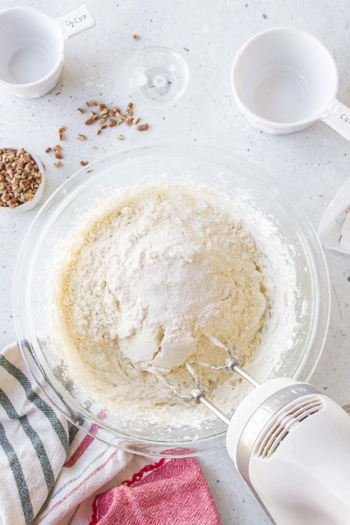 a clear glass bowl with an electric mixer in it that has mixed powdered sugar, butter, and vanilla and about to mix flour into it, with a kitchen towel beside the bowl, a small empty white bowl, a tiny bowl of chopped pecans, a tiny glass bowl of salt and a measuring cup of flour