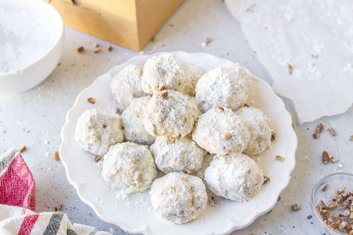 a white plate full of mexican wedding cookies sprinkled with chopped pecans with a brown box in the background, a kitchen towel beside the plate and a tiny bowl of chopped pecans and white bowl of powdered sugar around