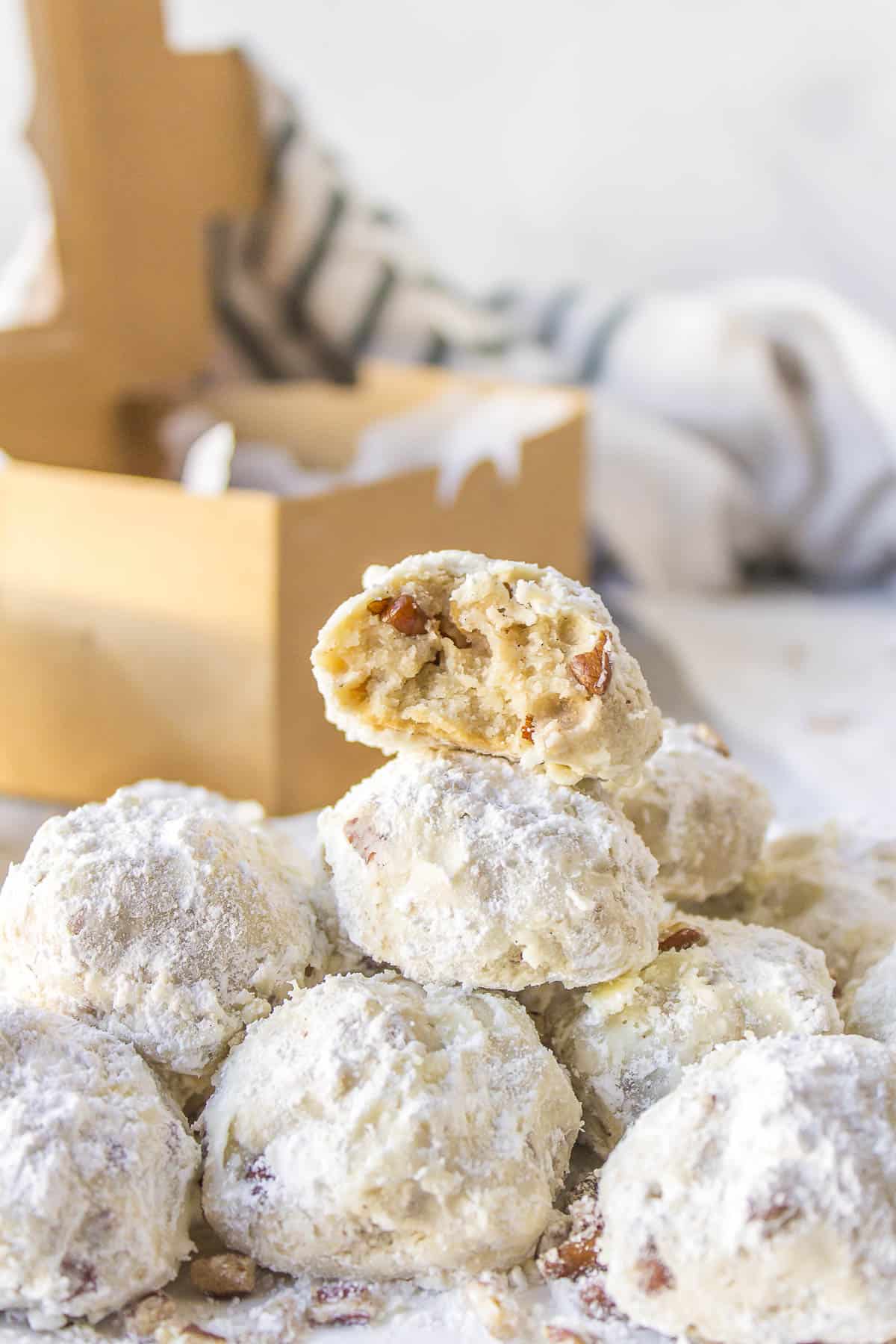 a pile of mexican wedding cookies with a bite out of the cookie on top of the pile, chopped pecans sprinkled around, and a brown box in the background