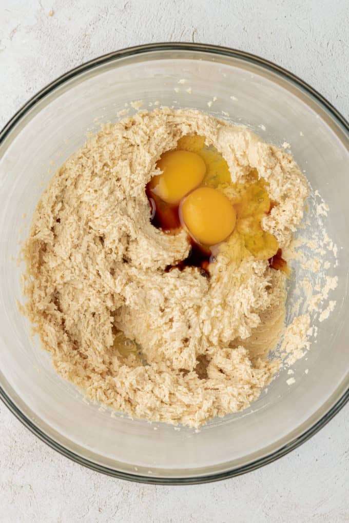 a clear glass bowl with mixed butter, granulated sugar and brown sugar with two eggs and vanilla extract on top