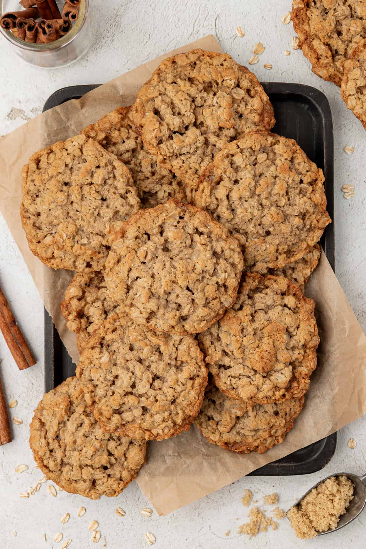 a sheet pan with parchment paper and a pile of oatmeal cookies on top with cinnamon sticks, more cookies and a spoon of brown sugar around it