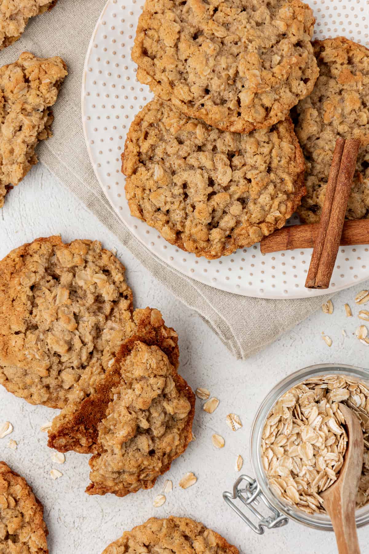 a plate of oatmeal cookies on top of a tan kitchen towel with more cookies around it, a jar of oats with a wooden spoon and oats sprinkled around