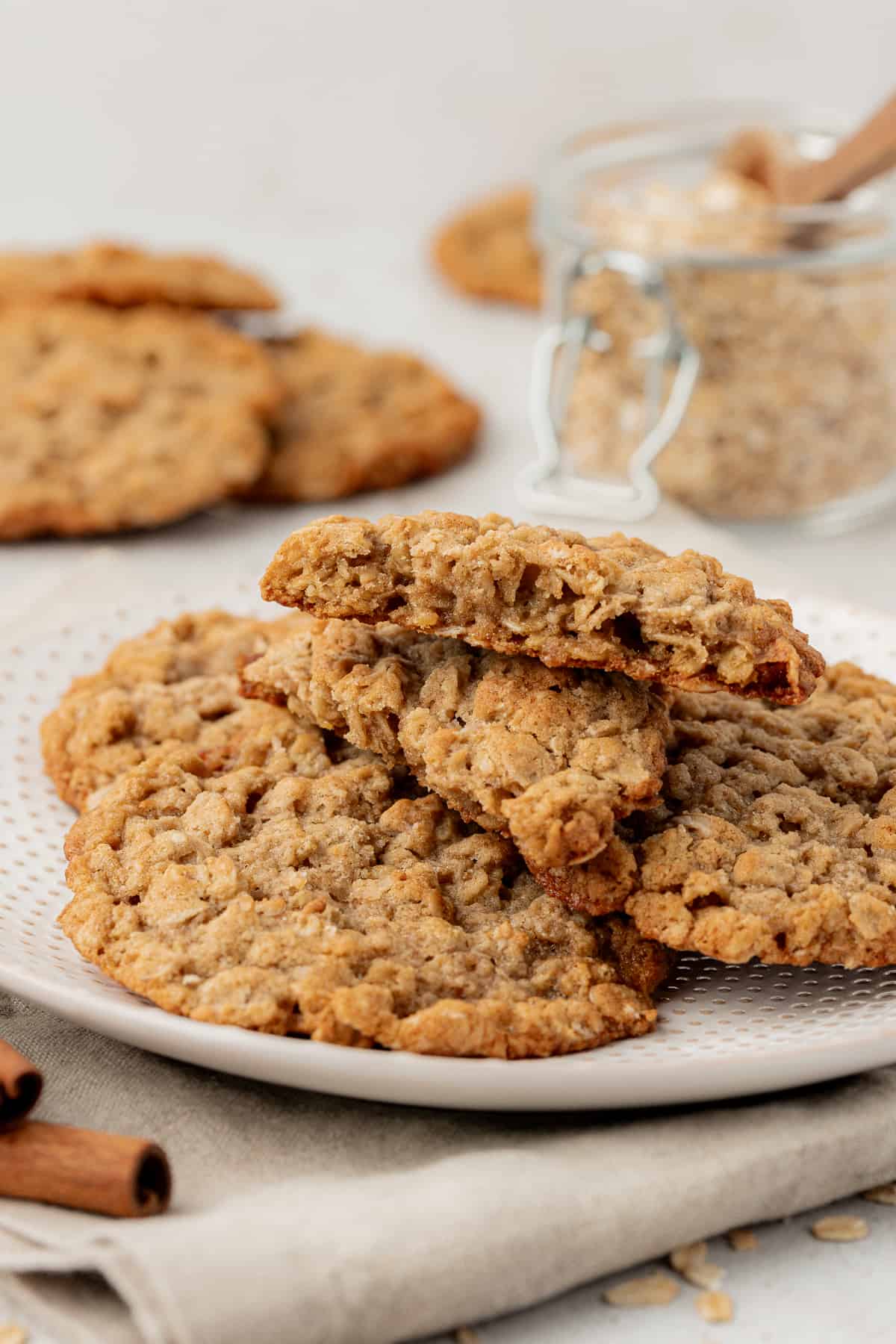 a white plate with a pile of oatmeal cookies on it on top of a tan kitchen towel with cinnamon sticks beside it, more cookies and a jar of brown sugar in the background