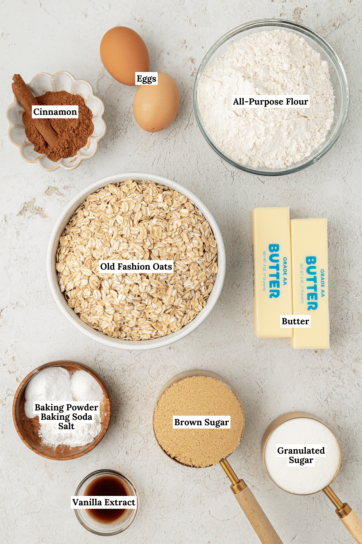 overhead view of oatmeal cookies ingredients including old fashioned oats, two sticks of butter, cinnamon, brown sugar, all-purpose flour, vanilla extract, two eggs, baking powder, baking sugar and salt