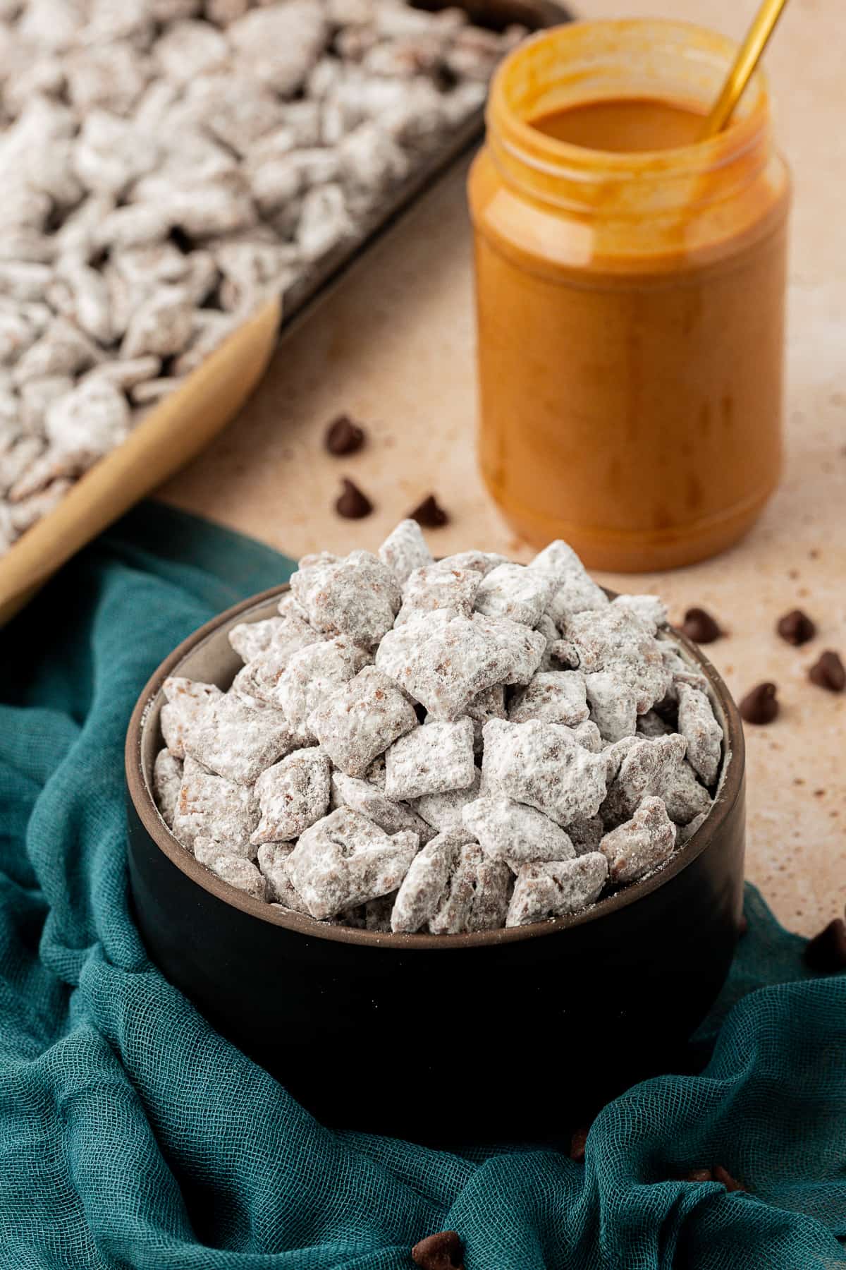 a black bowl full of puppy chow on a dark teal towel with a jar of peanut butter beside it and a sheet pan full of more puppy chow, with chocolate chips sprinkled around