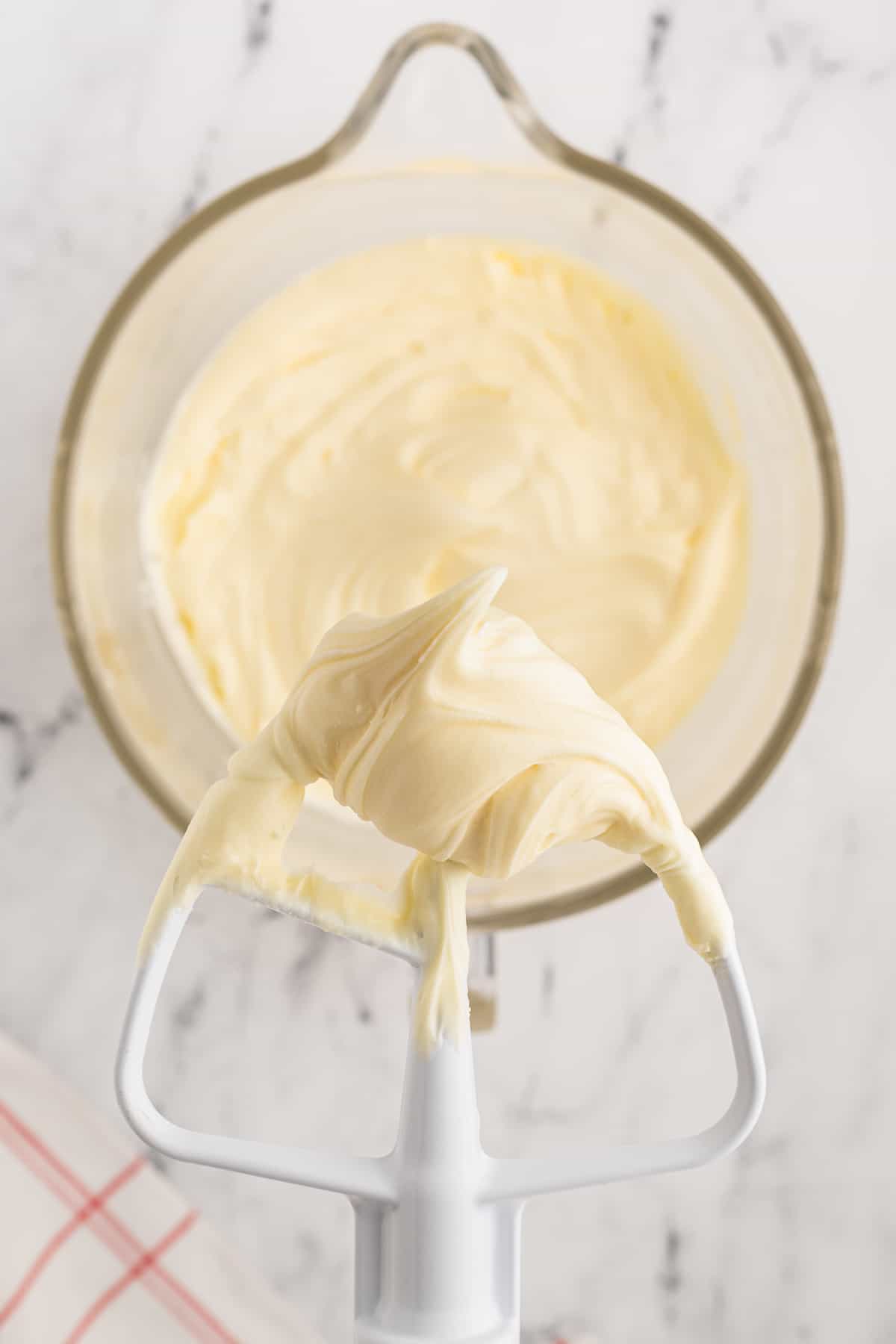 cream cheese frosting in a mixing bowl with the mixer lifted above it, covered in cream cheese frosting