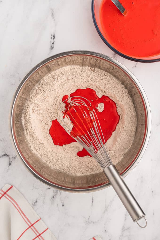dry ingredients for red velvet cake with red wet ingredients being poured in and mixed with a wire whisk in a silver bowl
