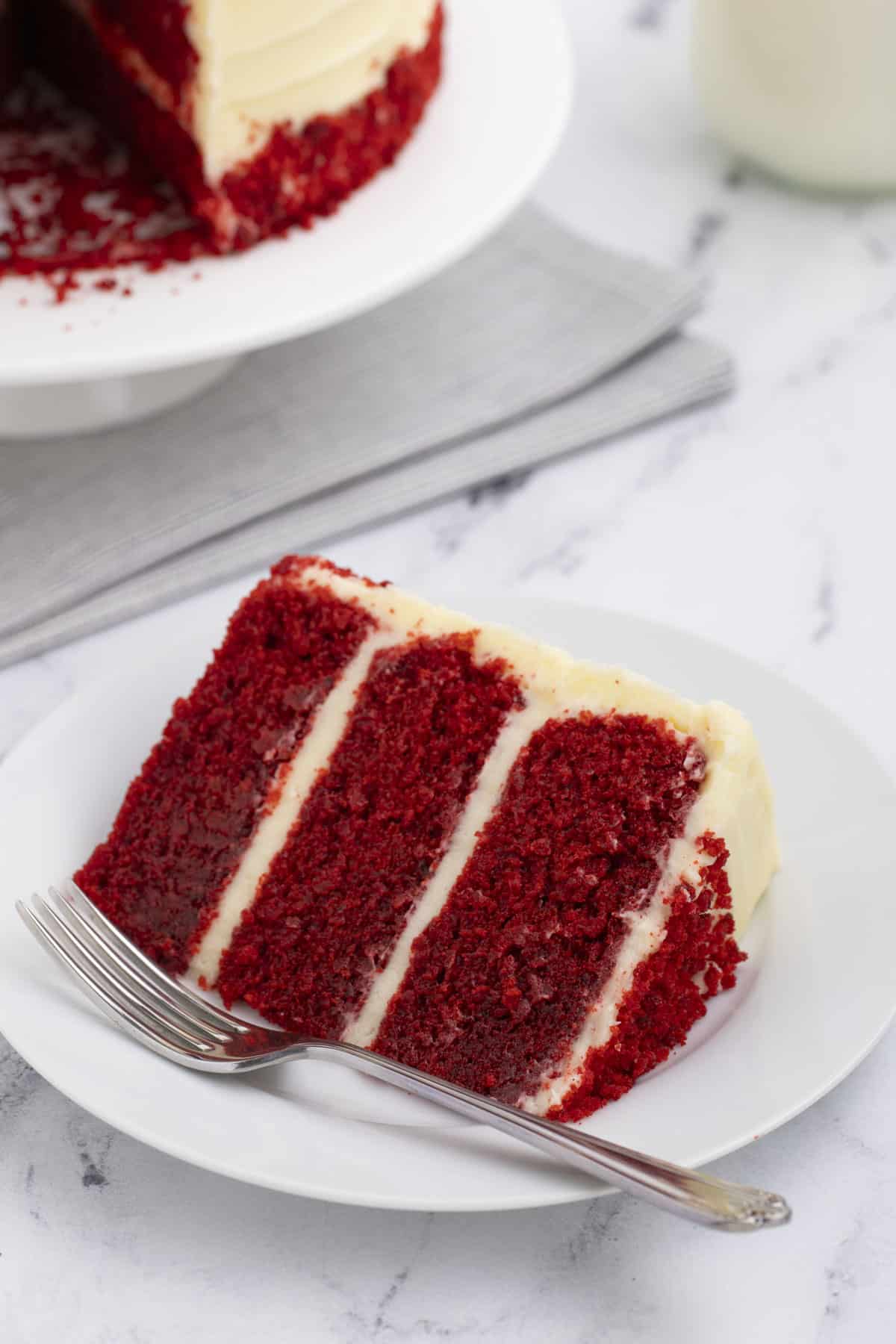 a slice of red velvet cake on a small white plate with a fork with the rest of the cake behind it on a white plate on top of a grey towel
