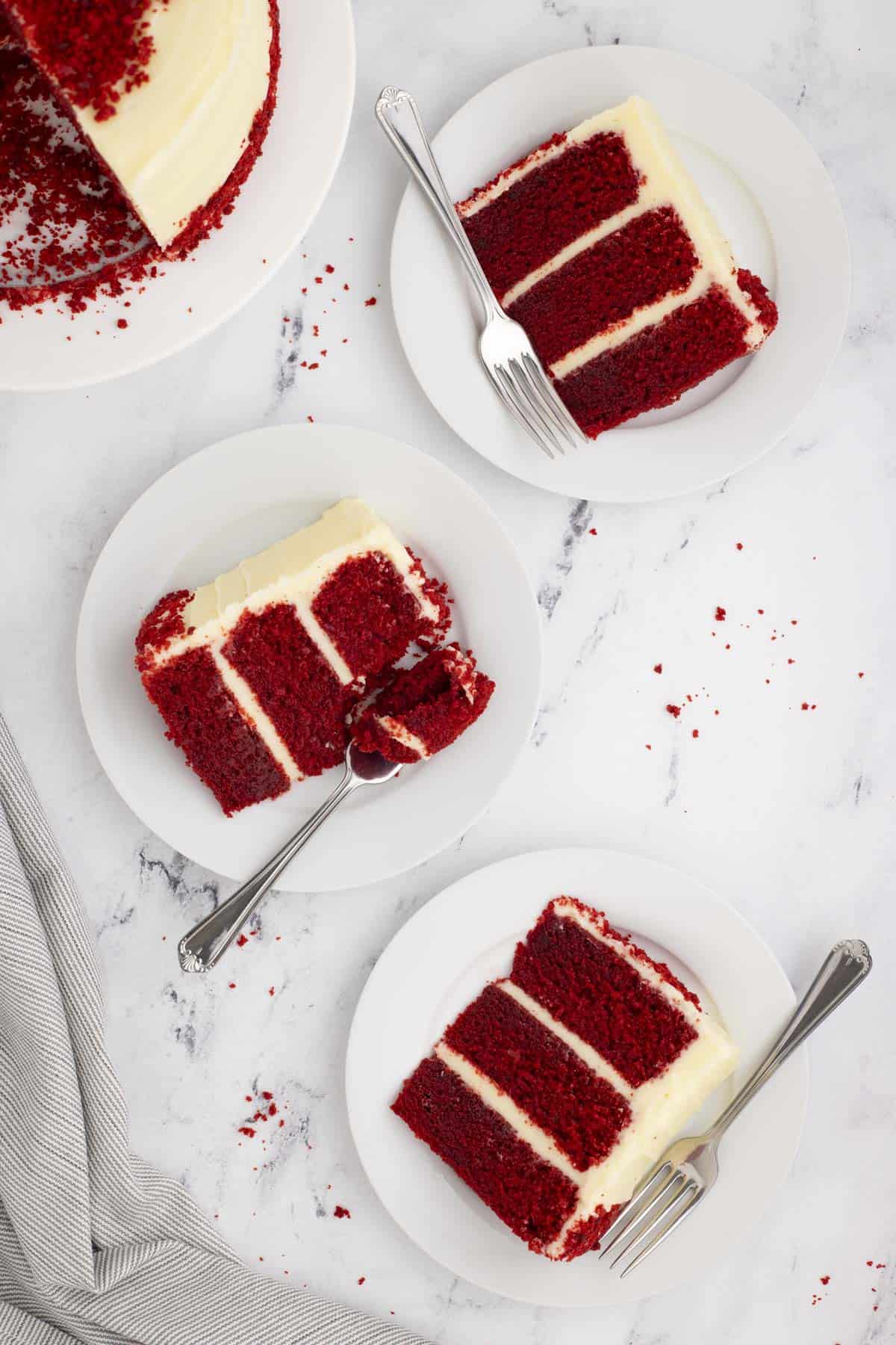 the overhead view of three slices of red velvet cake on white plates with forks beside another white plate with the remainder of the cake on it