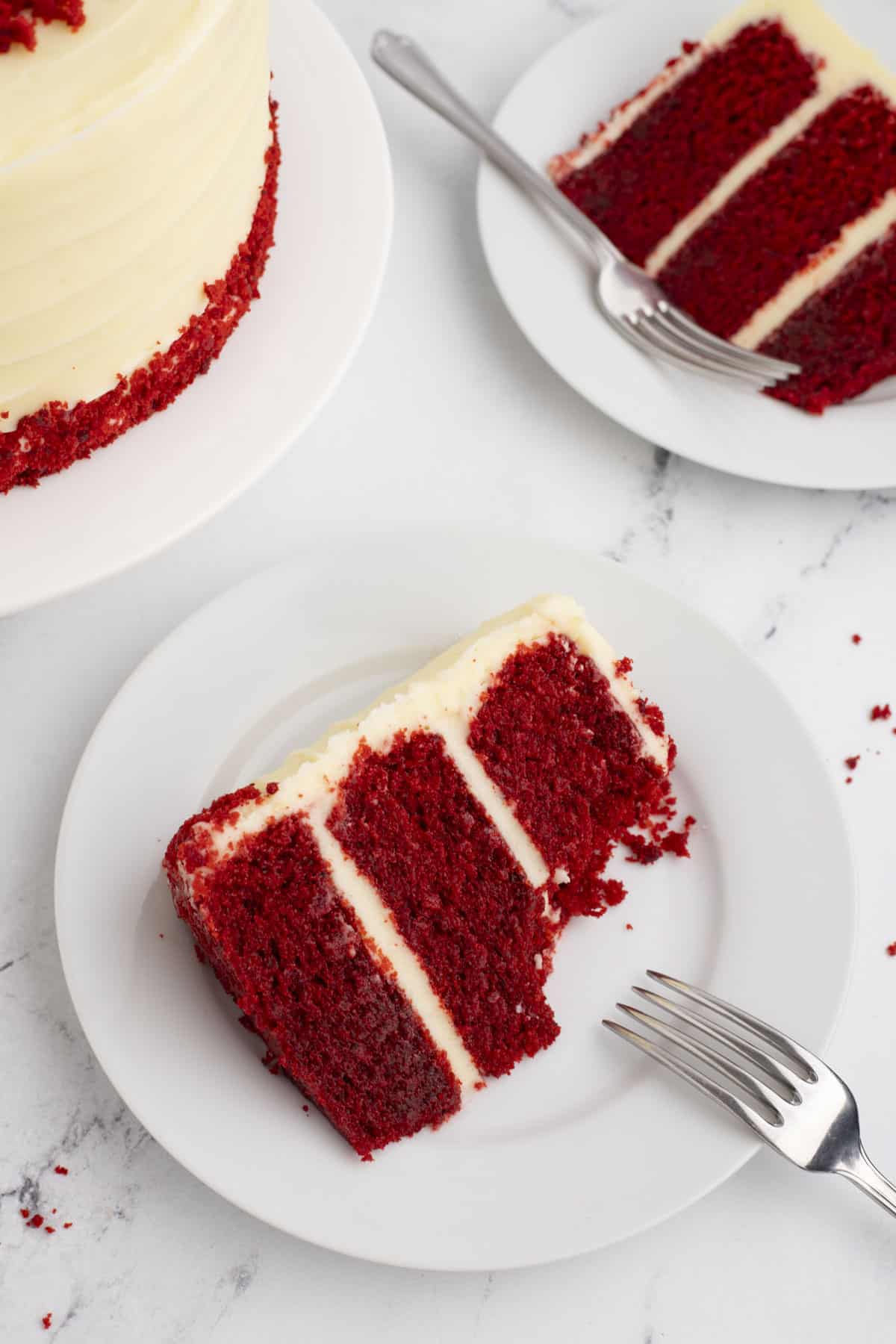 a slice of layered red velvet cake on a white plate with a fork leaning on it and another plate with a slice of cake and the edge of the whole cake in the background
