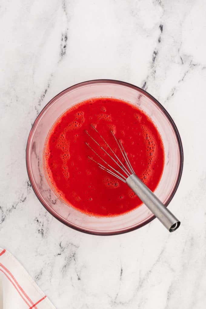 wet ingredients with bright red food coloring in it in a clear glass bowl with a wire whisk