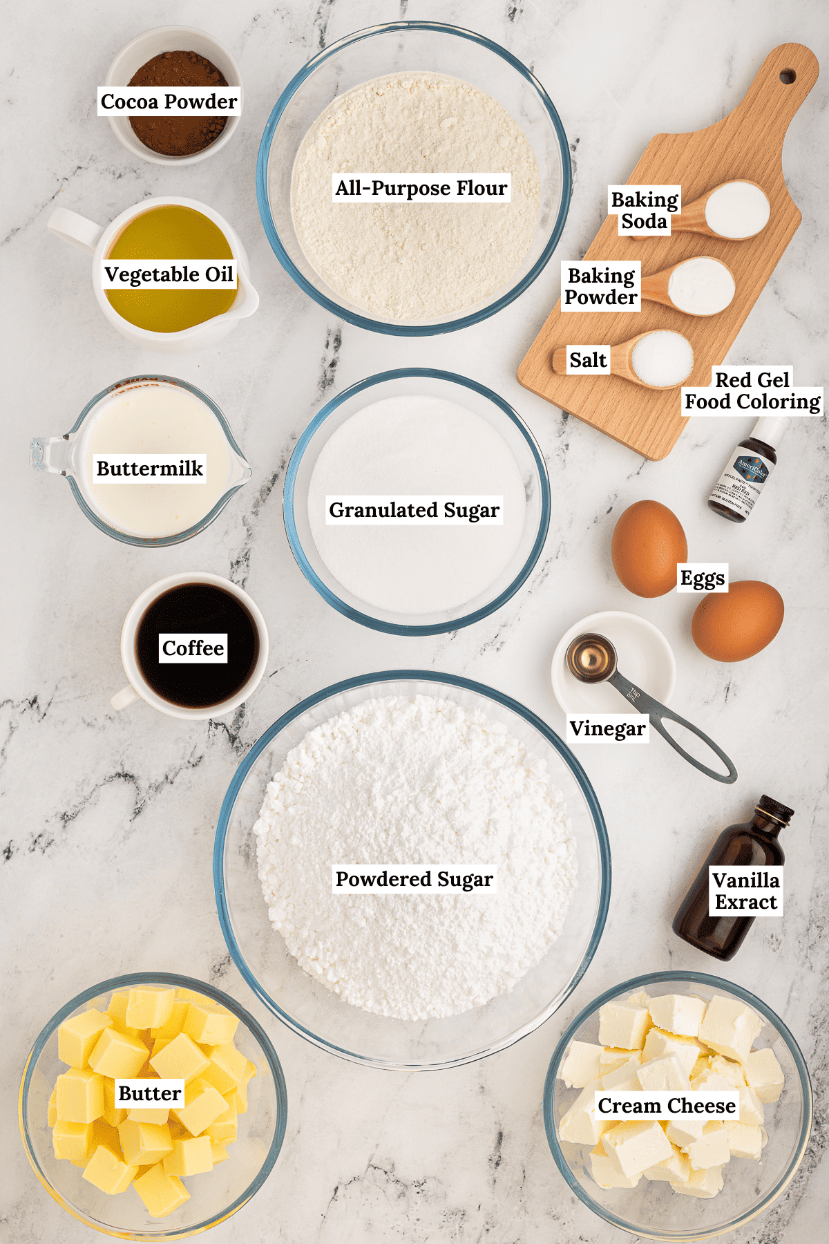 ingredients for red velvet cake arranged around each other on a counter including All-purpose flour, Dutch-processed cocoa powder, Baking soda, Baking powder, Salt, Granulated sugar, Eggs, Buttermilk, Vegetable oil, Vanilla extract, Red food coloring, Distilled vinegar, Instant coffee granules, Unsalted butter, Powdered sugar, Vanilla extract, and Cream cheese
