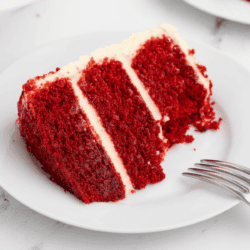 a slice of layered red velvet cake with cream cheese frosting on a white plate with a fork leaning on it