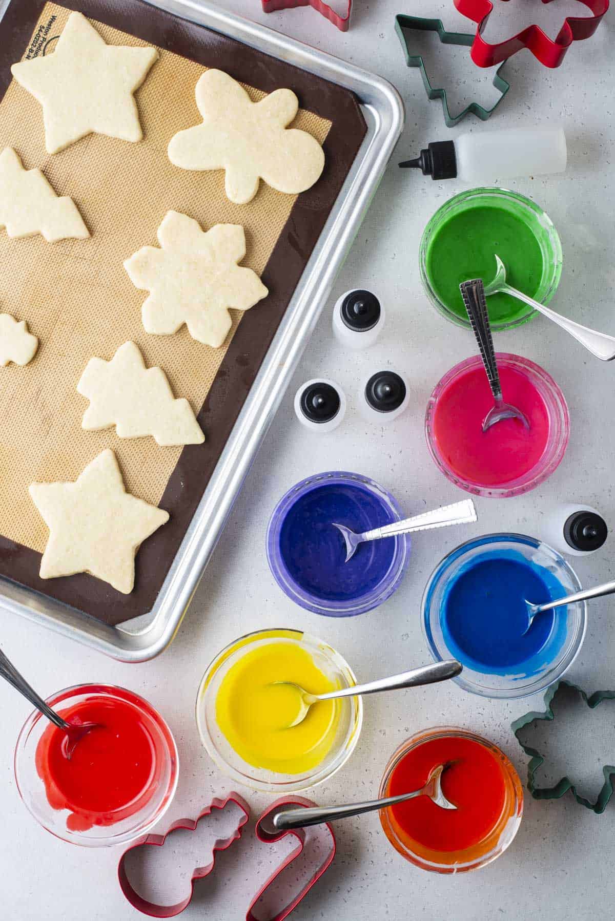 christmas shaped cookies lined in rows on a cookie sheet with bowls of colored icing with spoons beside it, empty squeeze bottles and cookie cutters