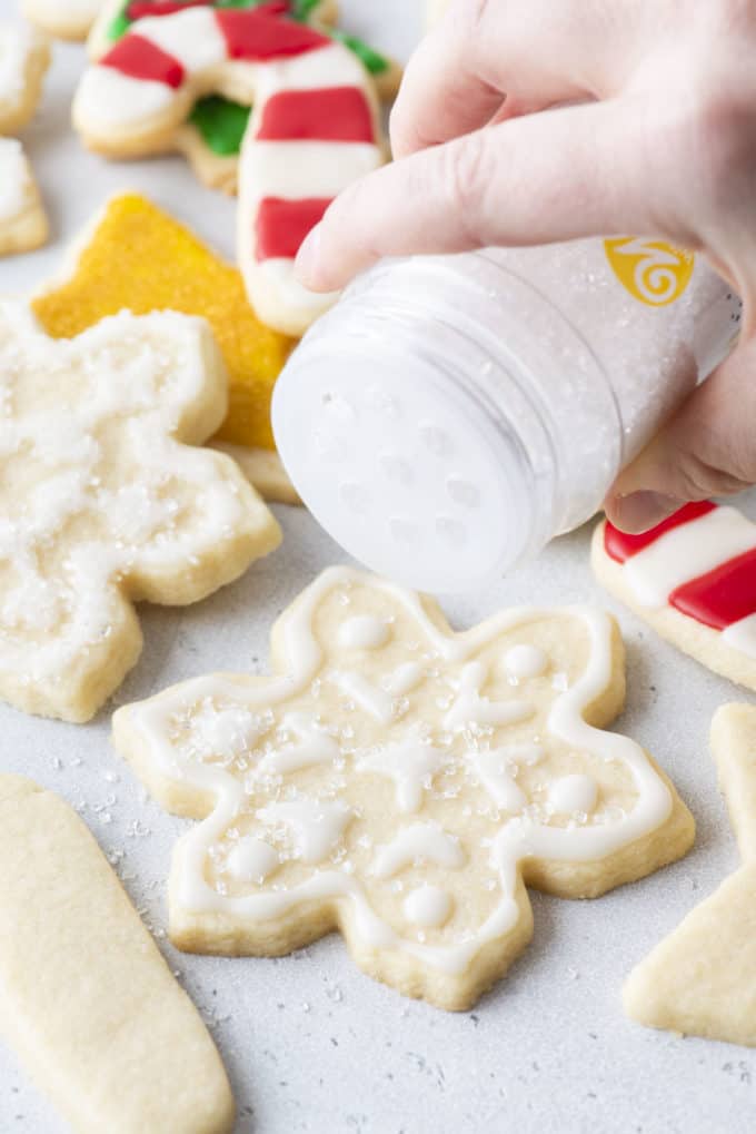 snowflake cookies with white royal icing being dusted with sugar with more christmas themed cookies in the background
