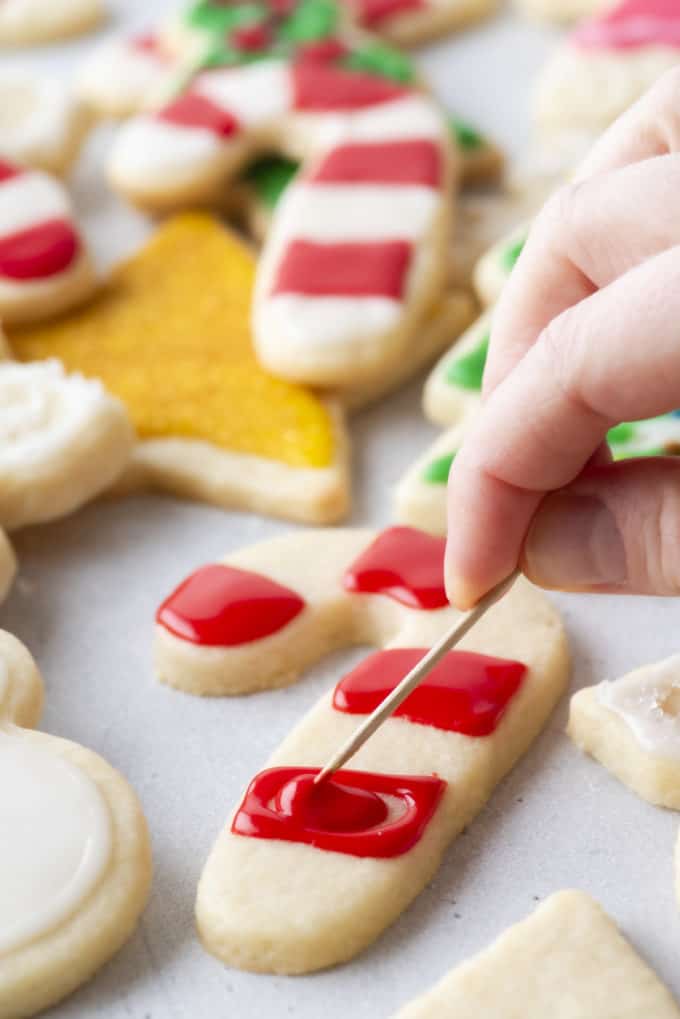 red royal icing being spread using a toothpick on a candy cane shaped cookie with more christmas themed cookies in the background