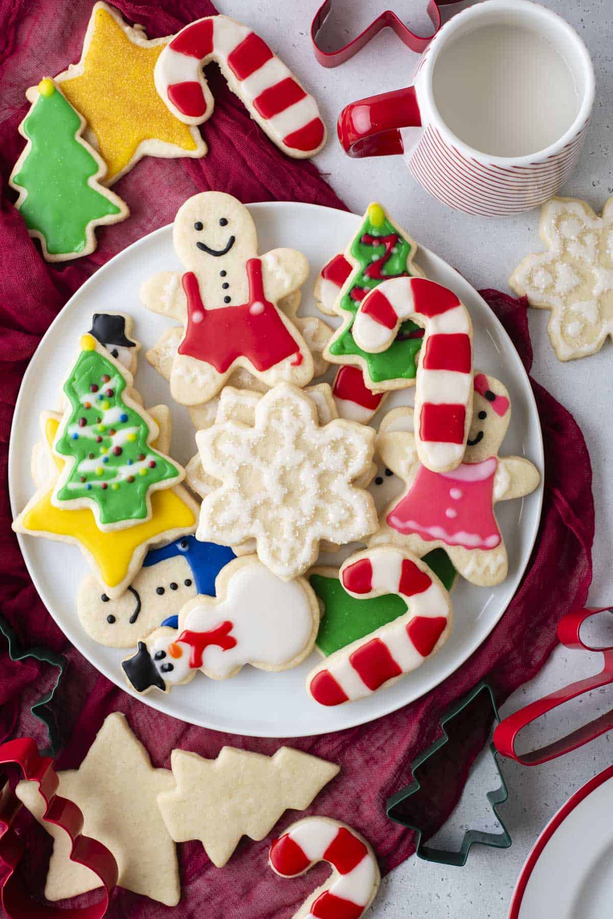 a plate full of christmas cookies decorate with royal icing on top of a red towel with more cookies, cookie cutters and a cup of milk around it