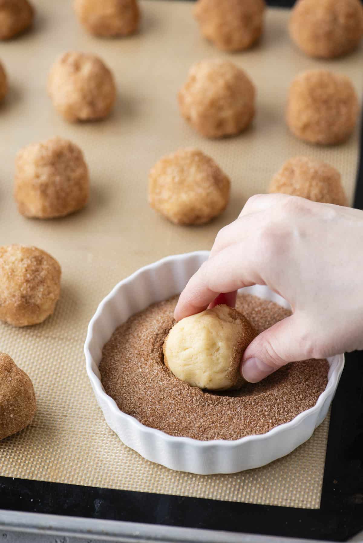 a hand dipping a cookie dough ball into a white dish of cinnamon sugar on a baking sheet lined with a silicone baking mat with rows of cookie dough balls on it