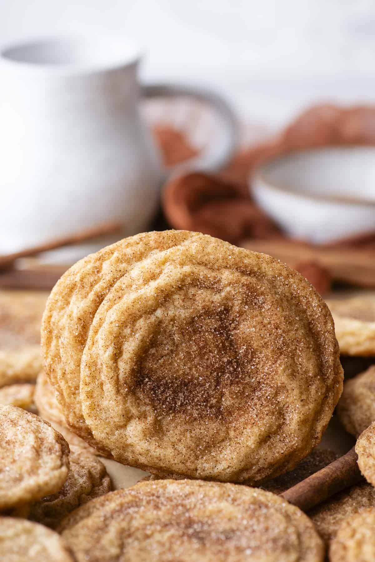 a snickerdoodle cookie standing straight up on its side surrounded by more cookies laying flat and a cinnamon stick, with a white cup of milk and bowl in the background
