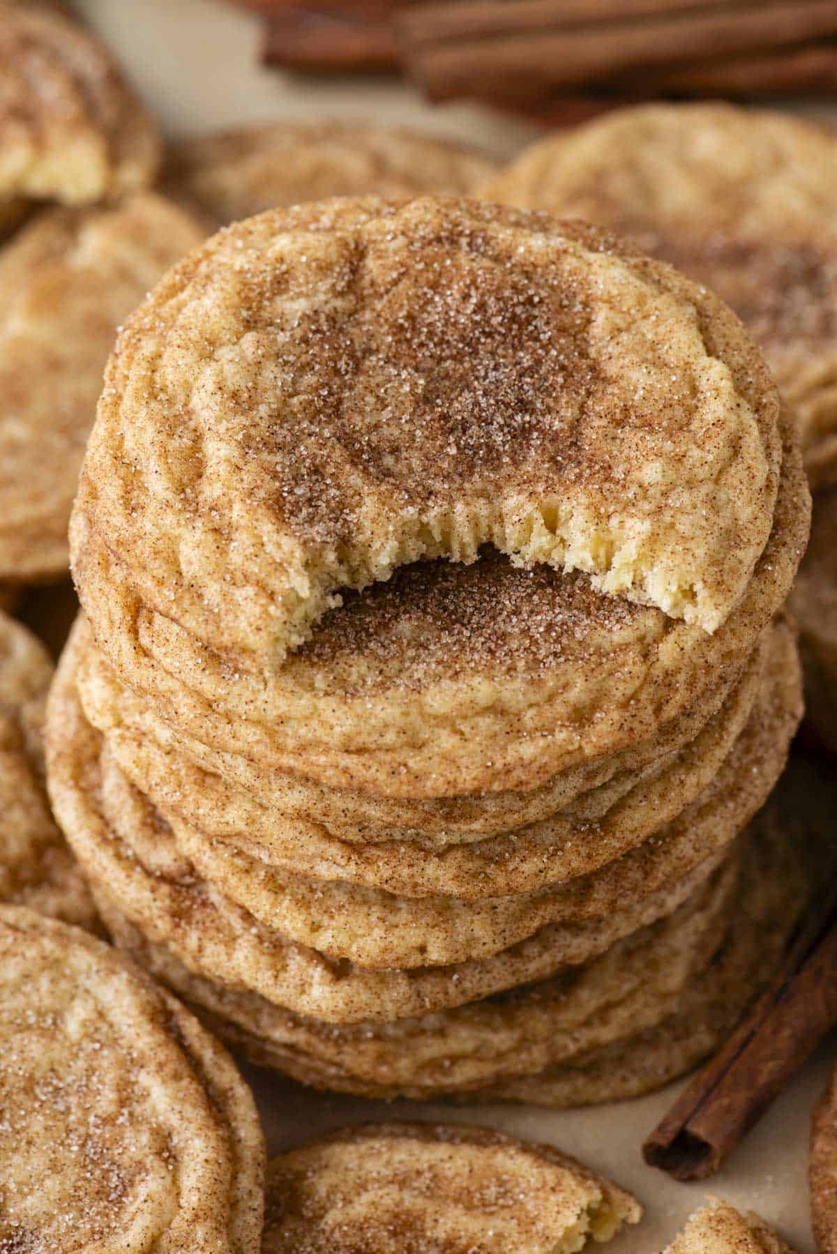 a stack of snickerdoodle cookies with a bite taken out of the cookie on top, and more cookies and a cinnamon stick surrounding it