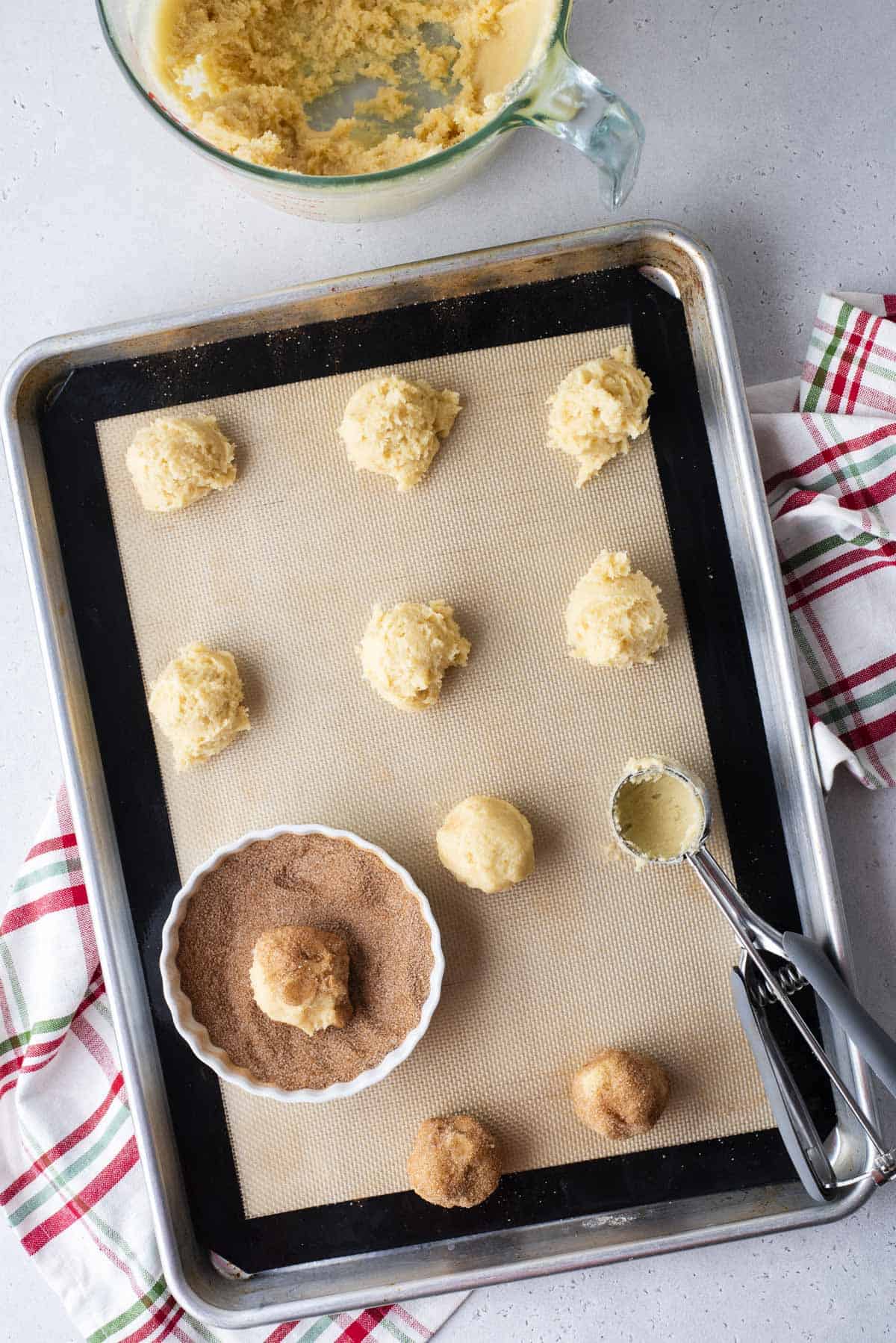 a sheet pan lined with a silicone baking mat with balls of snickerdoodle cookie dough in rows on it, with a cookie scoop and bowl of cinnamon sugar on the baking sheet, and two of the dough balls coated in the mixture. Beside the pan, a glass measuring dish with remnants of cookie dough in it and under the pan, a green, red and white plaid kitchen towel