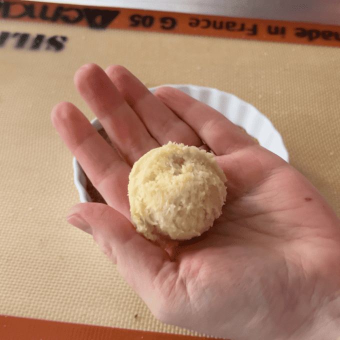 a hand hold a cookie dough ball flat on its palm over top of a white dish sitting on a silicone baking mat