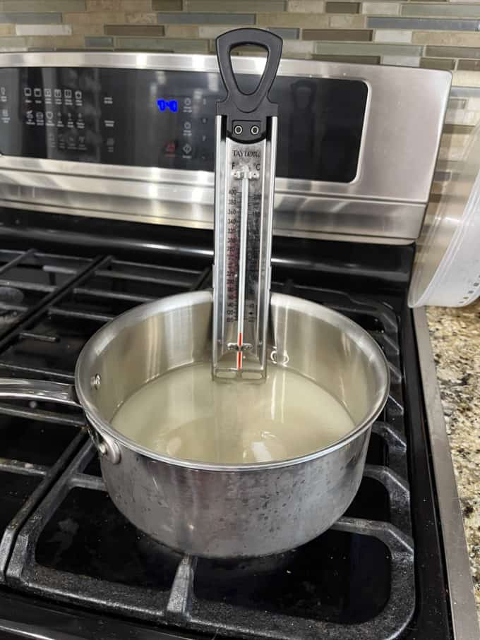 a pot on a stovetop with granulated sugar, water and corn syrup mixed in it and candy thermometer attached to the side
